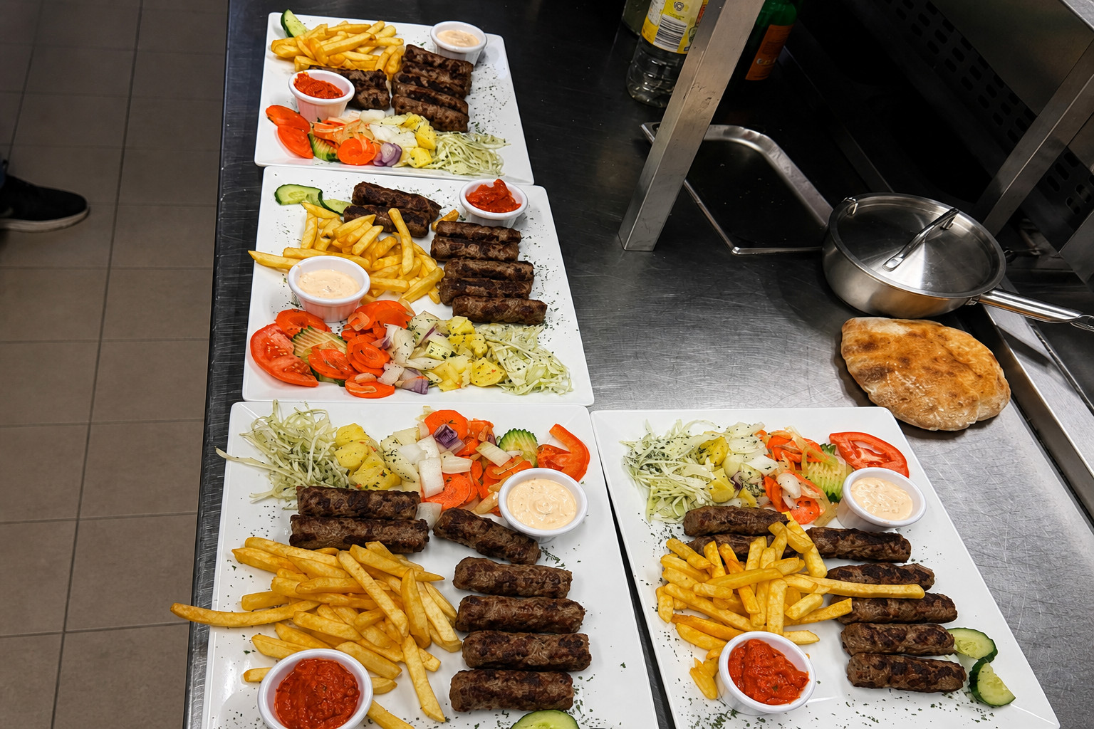 Mehrere Ćevapčići-Teller mit Pommes und Salat, frisch angerichtet in der Küche – Balkan Grill Pnishi